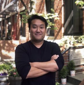 Daniel Yoo crosses his arms while posing for the photo. Behind him is a brick building covered in plants.