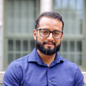 Sebastian Castillo-Hair smiles for the photo. He is wearing a blue collared shirt and glasses, and windows can be seen in the background.
